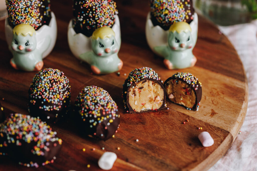 Peanut Butter Chocolate Eggs are on a wooden board. They are covered in chocolate and sprinkles. Three are sitting on the left edge of frame. One is cut open showing the peanut butter and marshmallow filling. Two of the eggs are in small chicken egg cups.