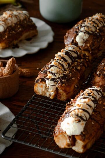 Chocolate banana cream buns are filled with whipped cream, drizzled with chocolate and crushed peanuts. 6 of them are on a black wire cooling rack. A whole one is in the back left corner on a ceramic plate. Another one can be seen in the front left corner. A wooden cup has whole peanuts in it, in the shells.