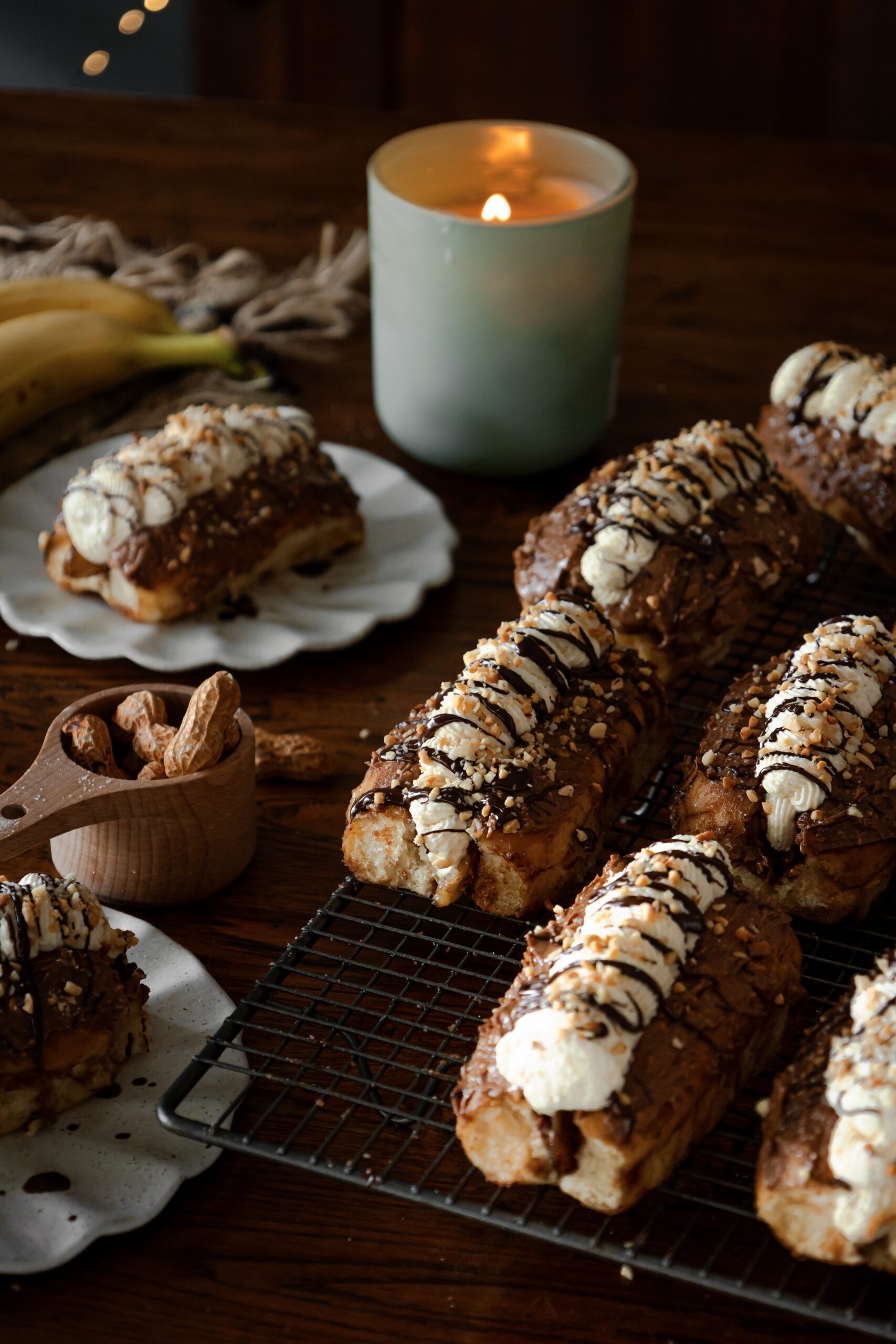 Chocolate banana cream buns are filled with whipped cream, drizzled with chocolate and crushed peanuts. 6 of them are on a black wire cooling rack. A whole one is in the back left corner on a ceramic plate. Another one can be seen in the front left corner. A wooden cup has whole peanuts in it, in the shells.