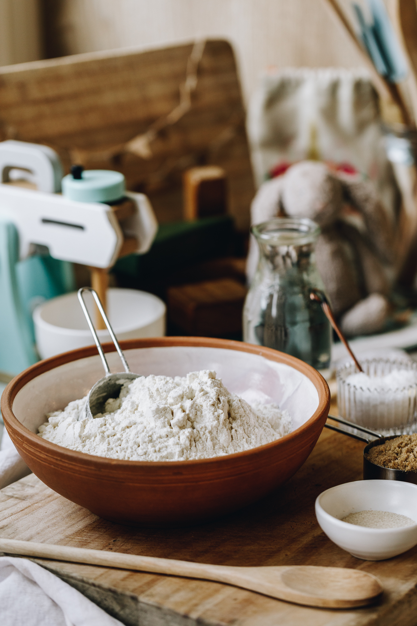A wooden board sits on white table cloth, on the board is a ceramic bowl with flour in it, a wooden spoon, brown sugar, salt and yeast. A wooden mixer is in the background with a fluffy rabbit and wooden board.