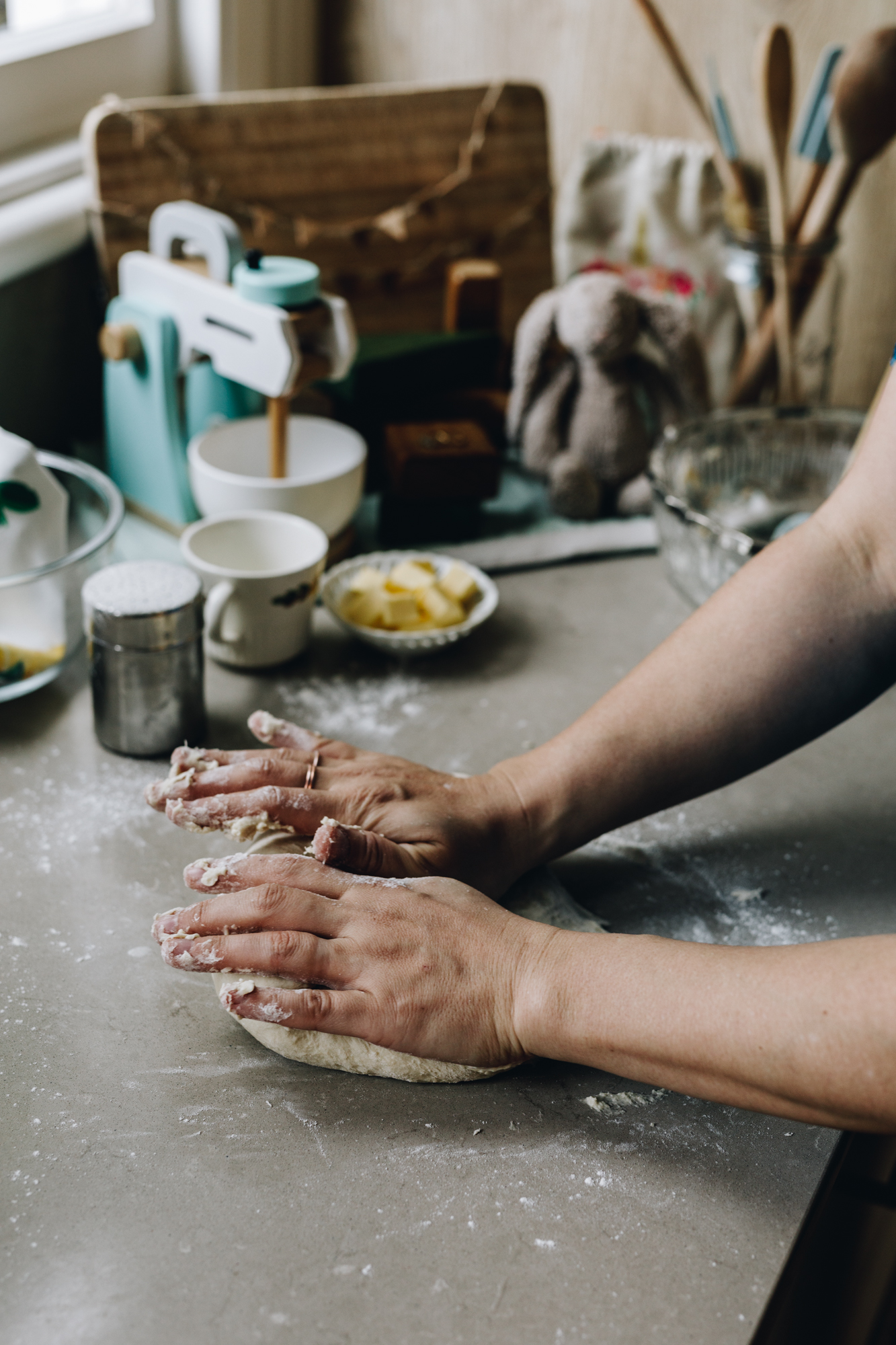 Naomi is kneading a dough on a floured grey bench. There is butter and cups. A wooden mixer is in the background with a fluffy rabbit and wooden board.