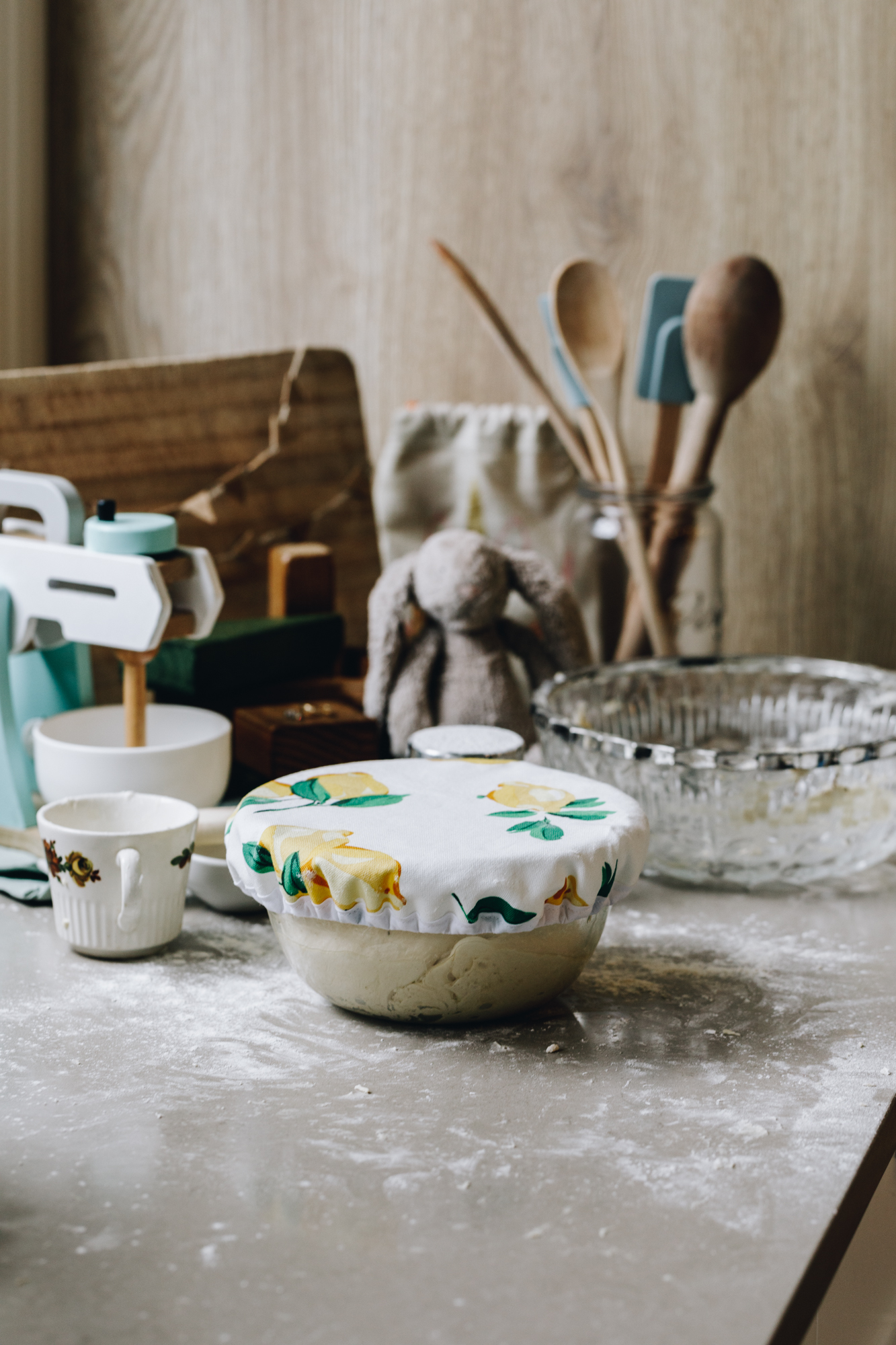 A glass bowl has a dough rising in it with a green and yellow lemon bowl cover. The bench is covered in dough with wooden spoons, a wooden kids mixer, a rabbit, a blow, cups and a wooden board in the background.