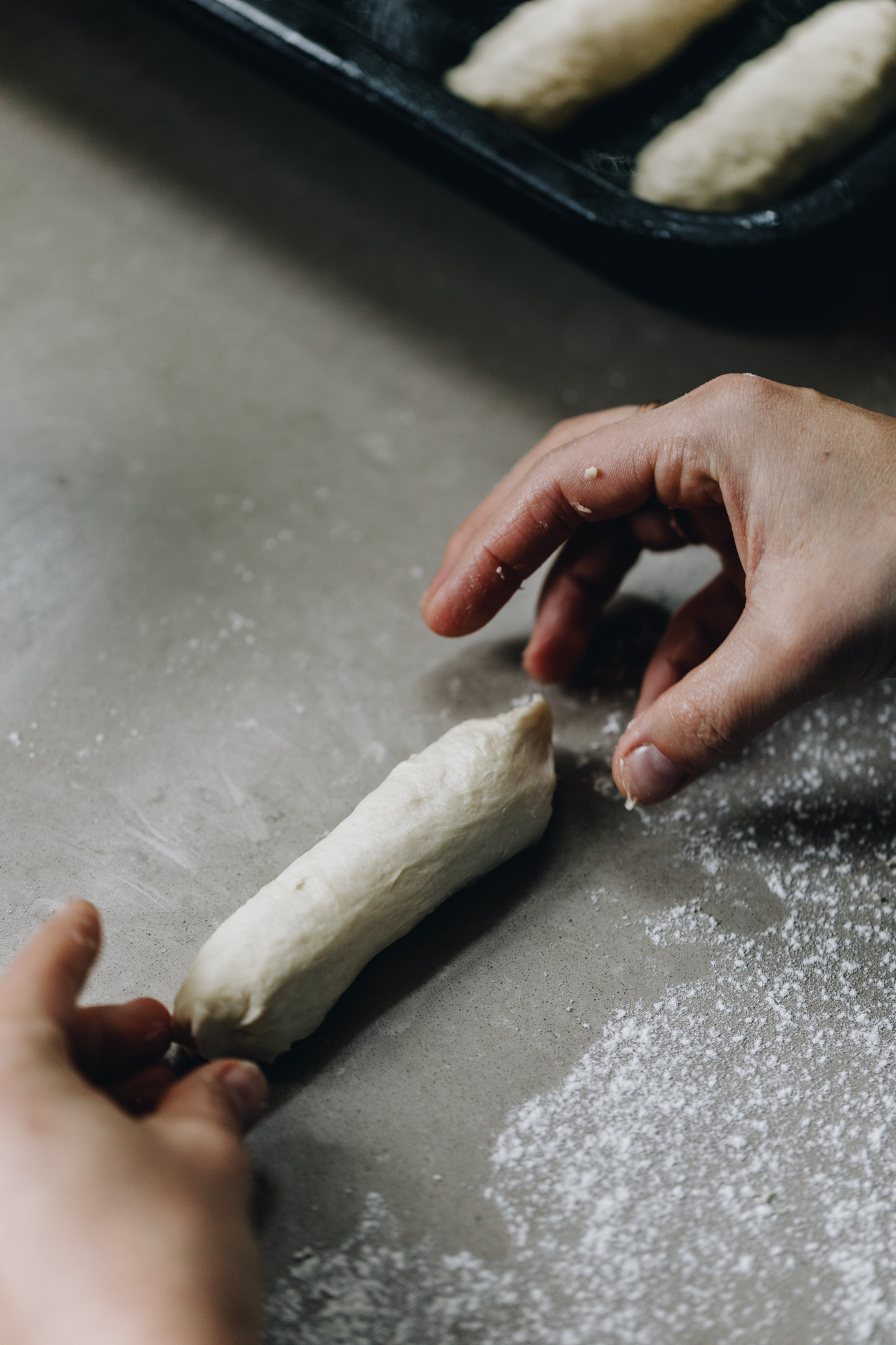 Naomi is shaping a dough in to a sausage shape on a great bench. Some of the buns are visible in a black tray in the corner of the shot.