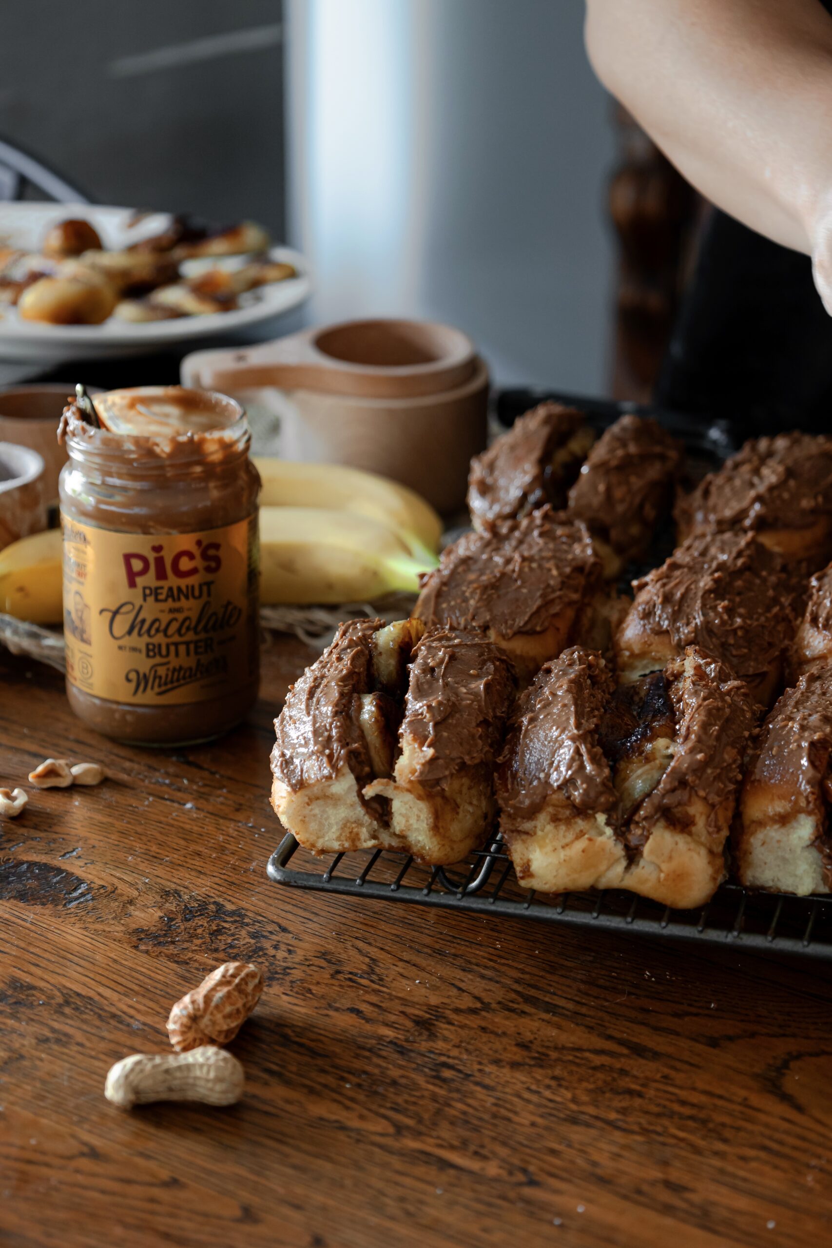 On a wooden bench, bread buns have been topped with chocolate peanut butter and filled with caramelised bananas. There is a jar of Pics Chocolate peanut butter and bananas with a wooden cup in the background.