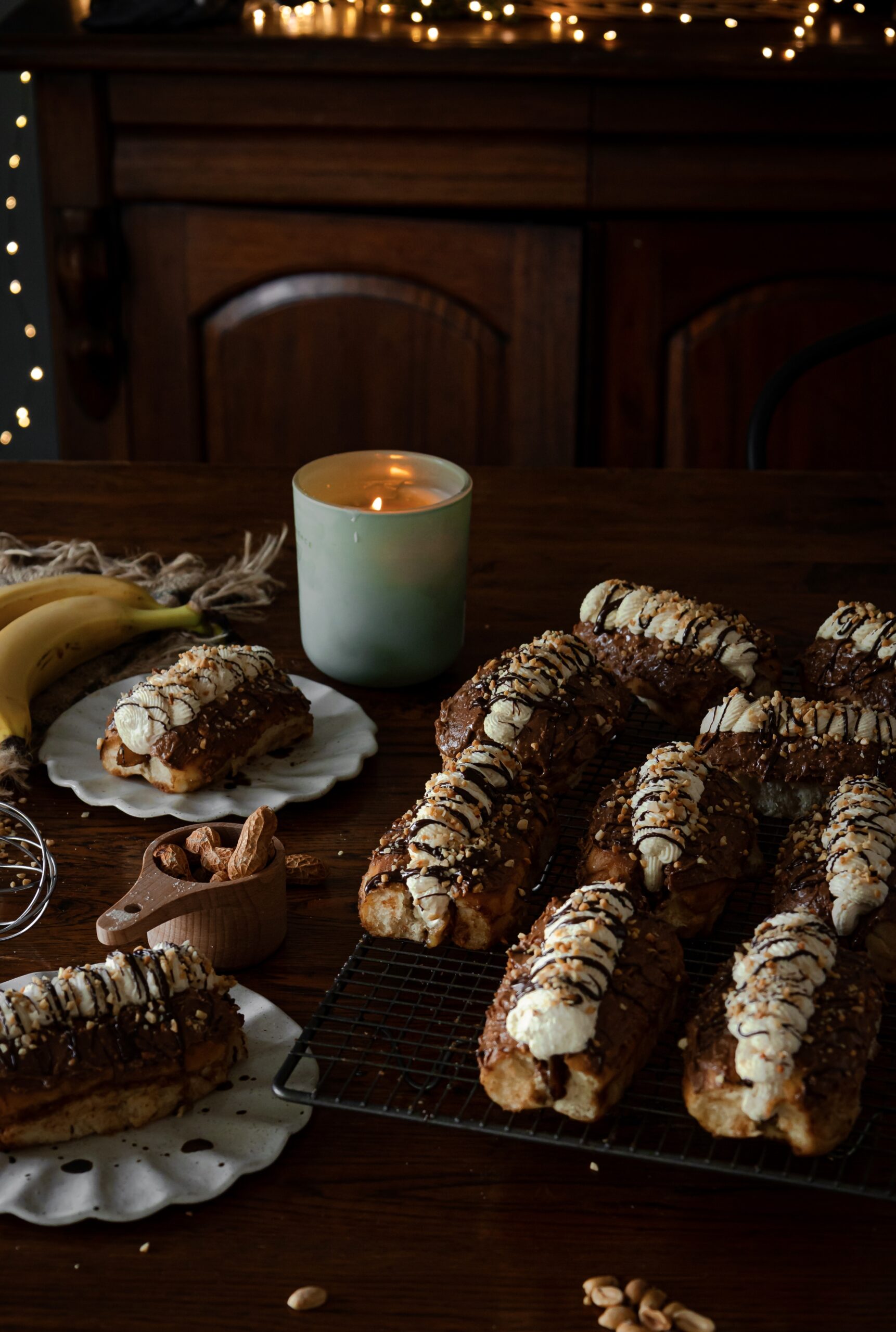 Chocolate banana cream buns are filled with whipped cream, drizzled with chocolate and crushed peanuts. 6 of them are on a black wire cooling rack. A whole one is in the back left corner on a ceramic plate. Another one can be seen in the front left corner. A wooden cup has whole peanuts in it, in the shells.