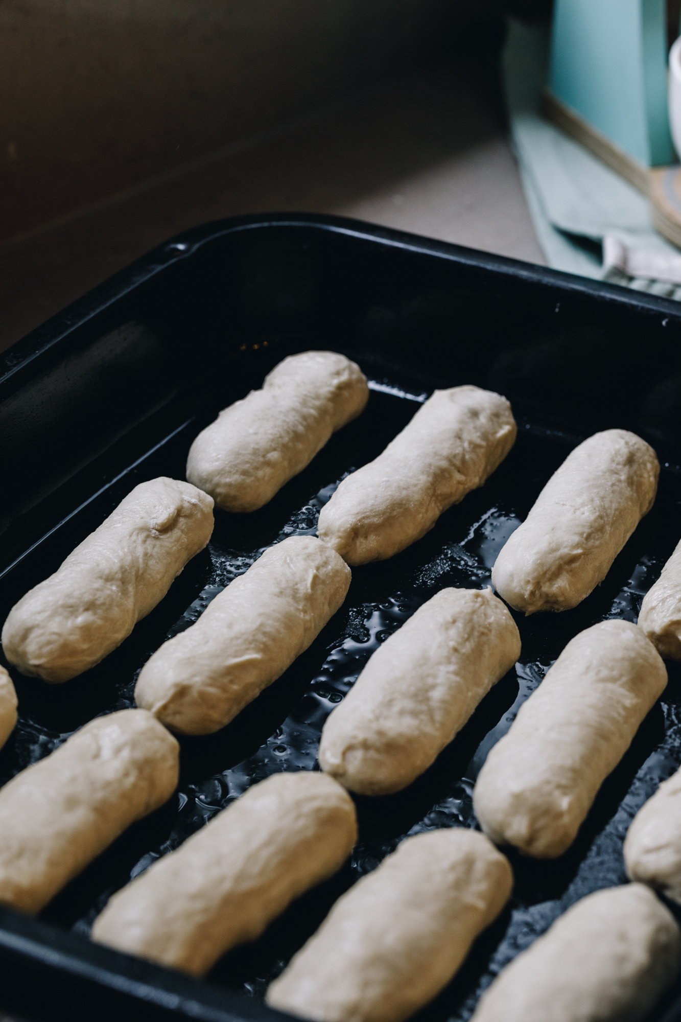 A greased tray has many sausage shaped buns in it.
