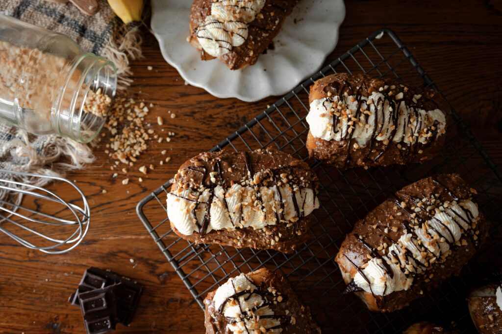 A top shot shows Chocolate banana cream buns are filled with whipped cream, drizzled with chocolate and crushed peanuts. 6 of them are on a black wire cooling rack. A whole one is in the back left corner on a ceramic plate. Another one can be seen in the front left corner.
