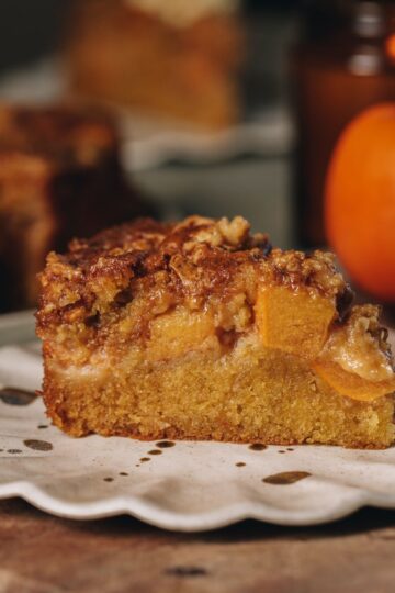 A slice of Persimmon Crumble Cake is on a a ruffled ceramic plate. It has a cake layer, chunks of persimmon and a crumble topping. There is a persimmon in the background.