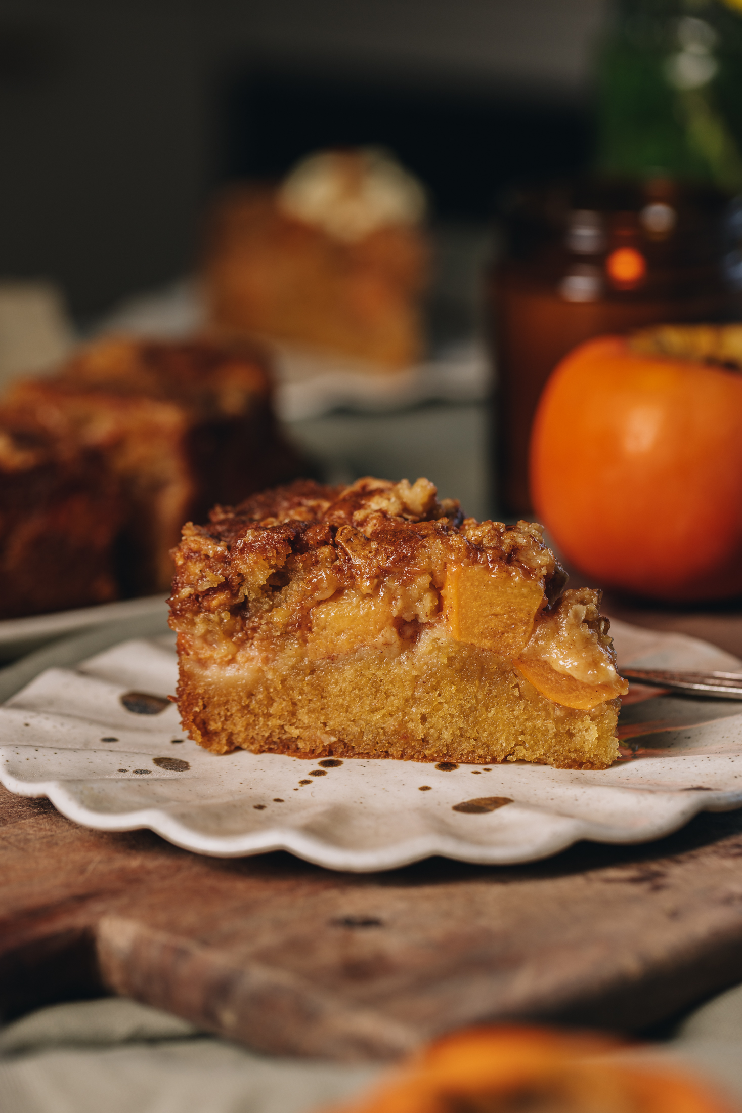 A slice of Persimmon Crumble Cake is on a a ruffled ceramic plate. It has a cake layer, chunks of persimmon and a crumble topping. There is a persimmon in the background.