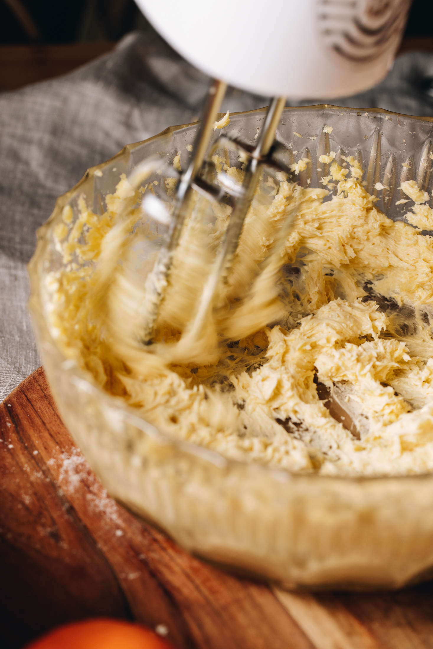 A glass vintage bowl is on a round wooden board on a grey tablecloth. Butter and sugar is being whipped with a white hand mixer. 