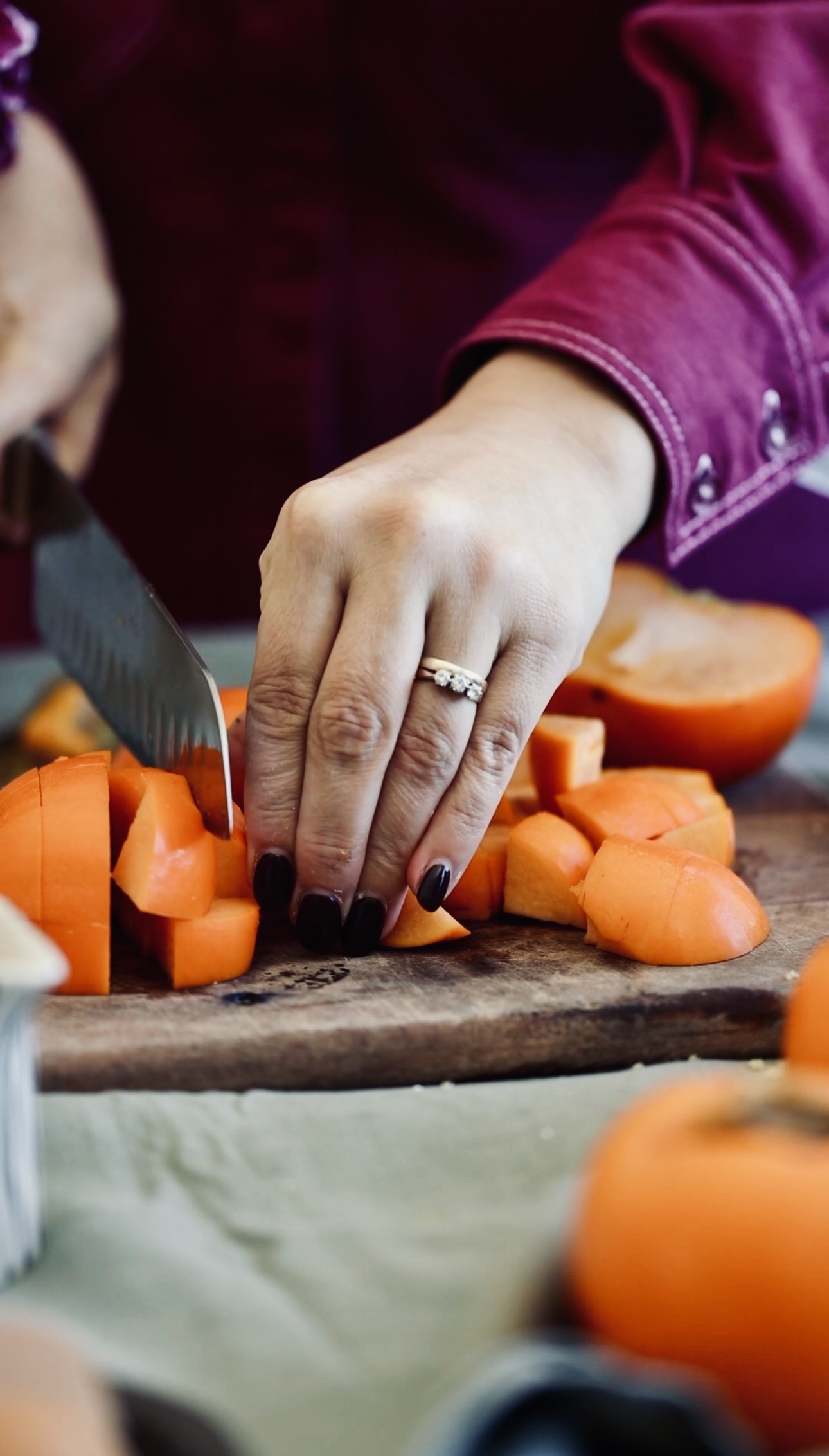 A wooden board sits on a green tablecloth. On the board is chunks of fresh persimmons. Naomi is cutting some more. 