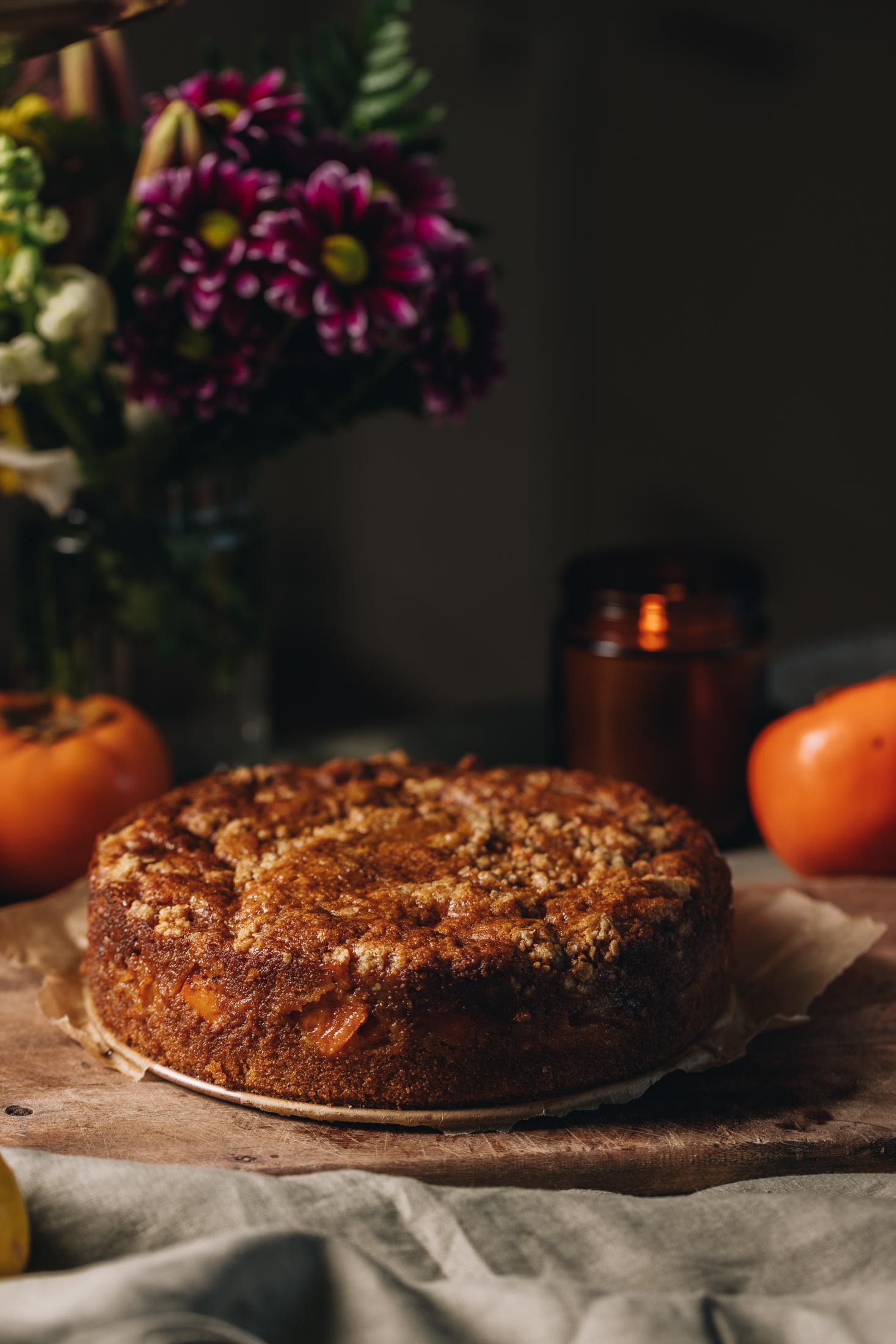 A persimmon crumble cake has been baked and is sitting on a wooden board. Persimmons are sitting behind it and a large bouquet of flowers are the left hand corner. 