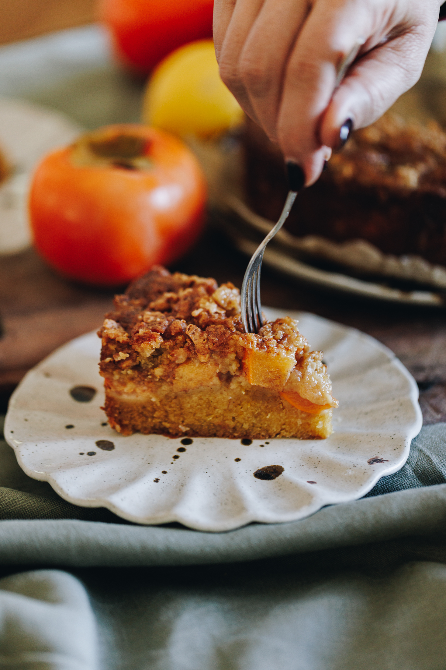 A slice of Persimmon Crumble Cake is on a a ruffled ceramic plate. It has a cake layer, chunks of persimmon and a crumble topping. Naomi is using a fork to take some of the cake. There is a persimmon in the background.