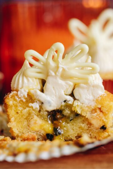 A white chocolate passionfruit muffins is cut open to reveal the gooey passionfruit centre. It is topped with a cream cheese frosting and a decoration. There are other muffins in the background.