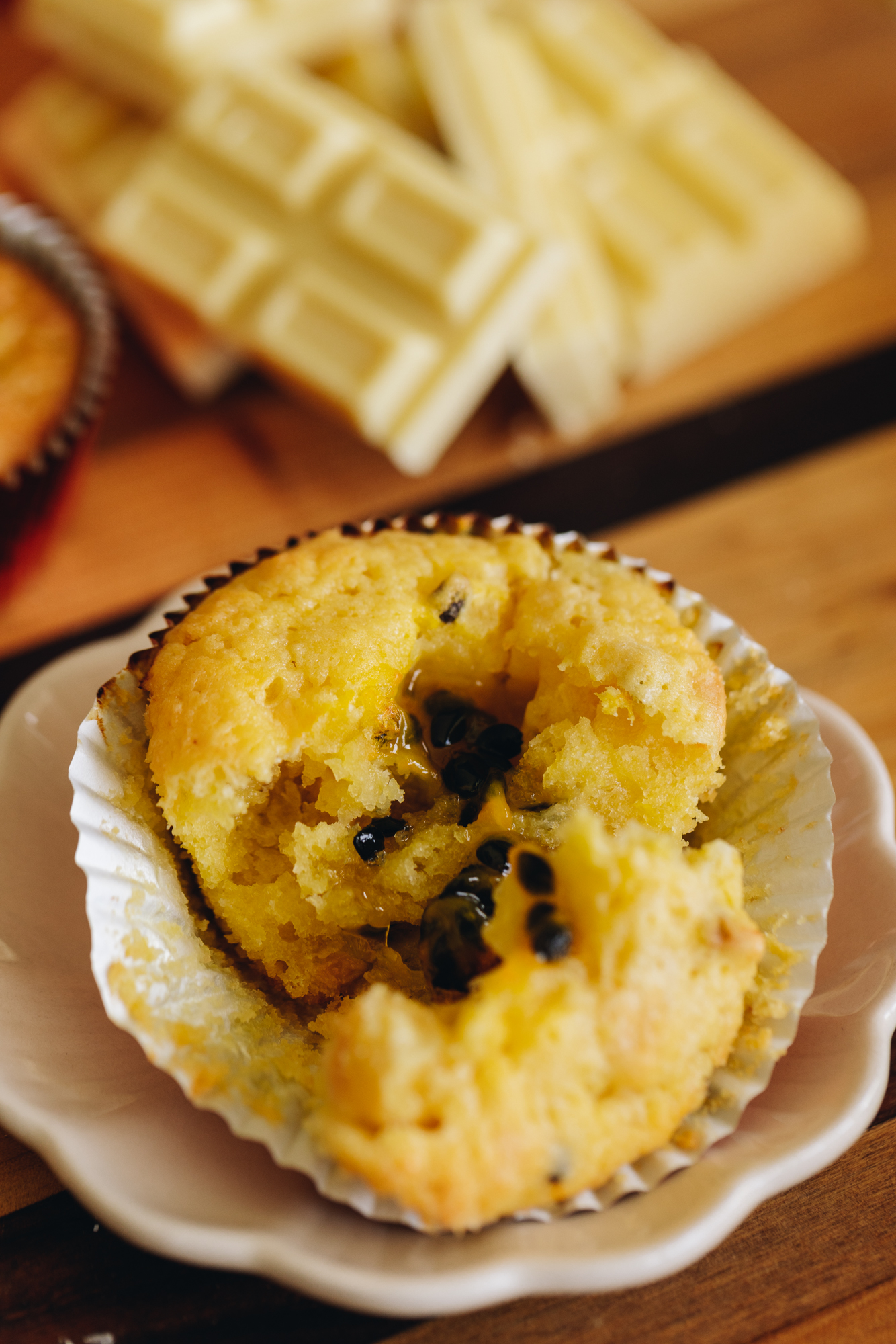 A white chocolate passionfruit muffin is broken open revealing the passionfruit filling. White chocolate is stacked behind it. 