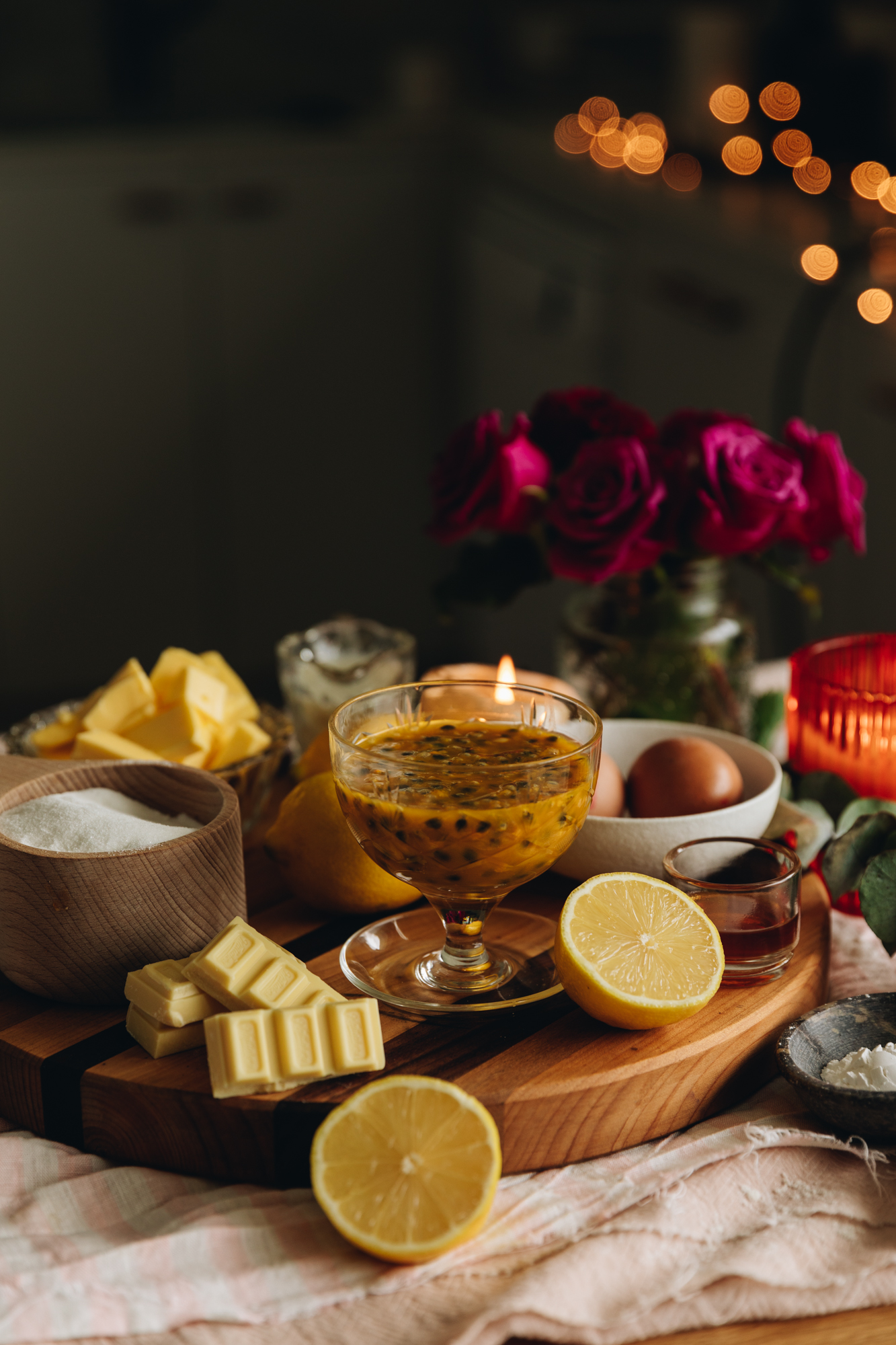 A striped board sits on a pink tablecloth is ingredients in different vintage vessels. There is sliced open lemons, passionfruit pulp, white chocolate, sugar, butter and a candle on the board. There is purple roses in a vase in the background. 