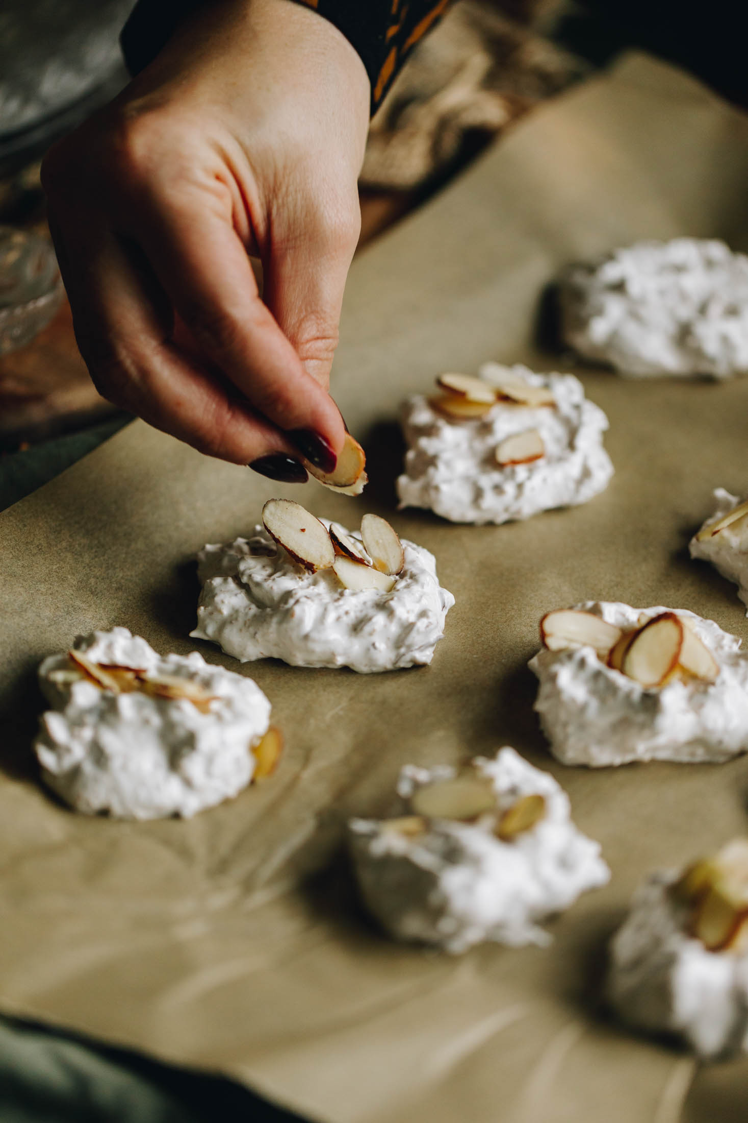 Meringue cookie mixture on a black tray lined with baking paper, with sliced almonds added on top of the cookies.