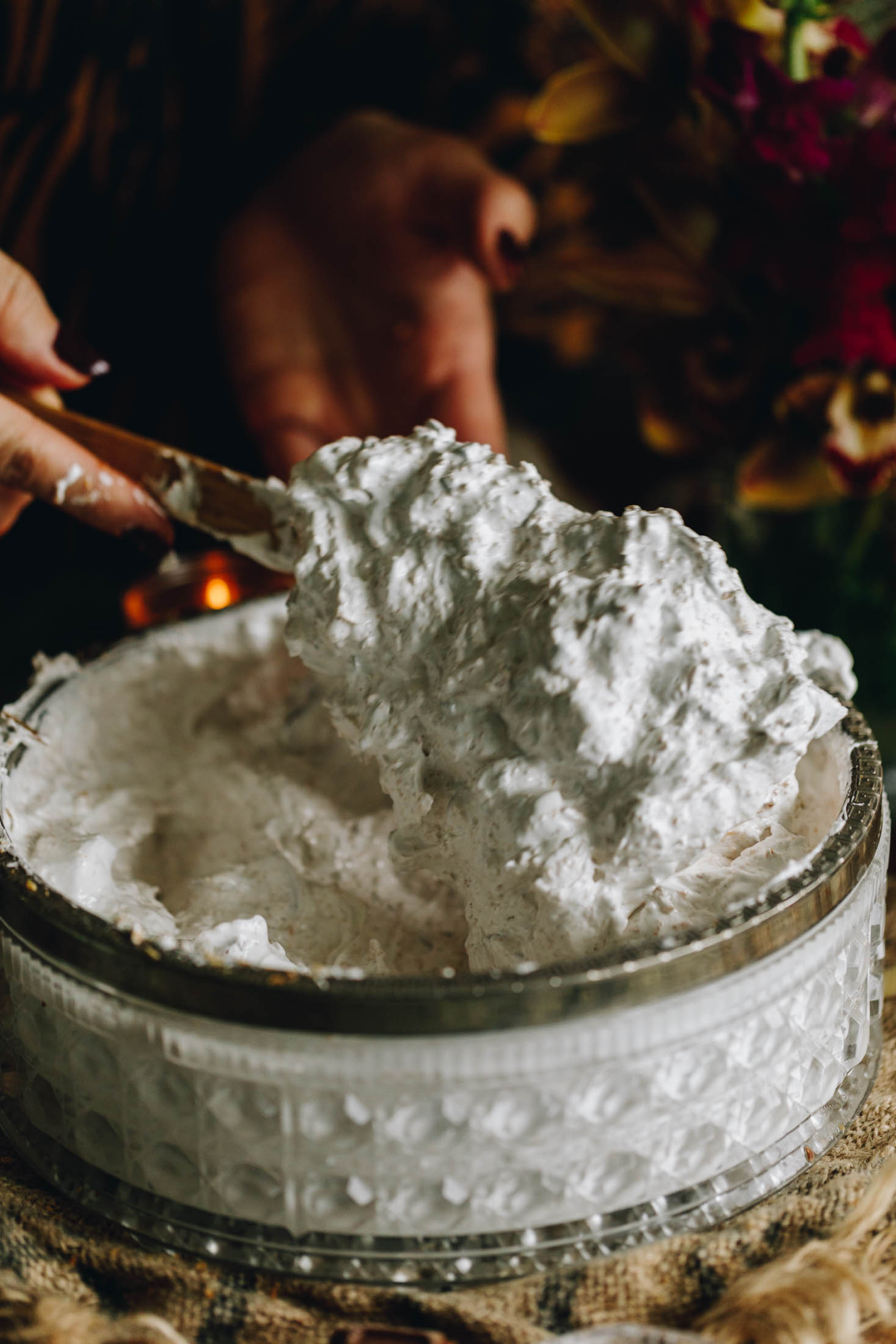 White meringue with coconut and chocolate in a vintage bowl on a wooden board, being stirred by hand with styling props in the background.