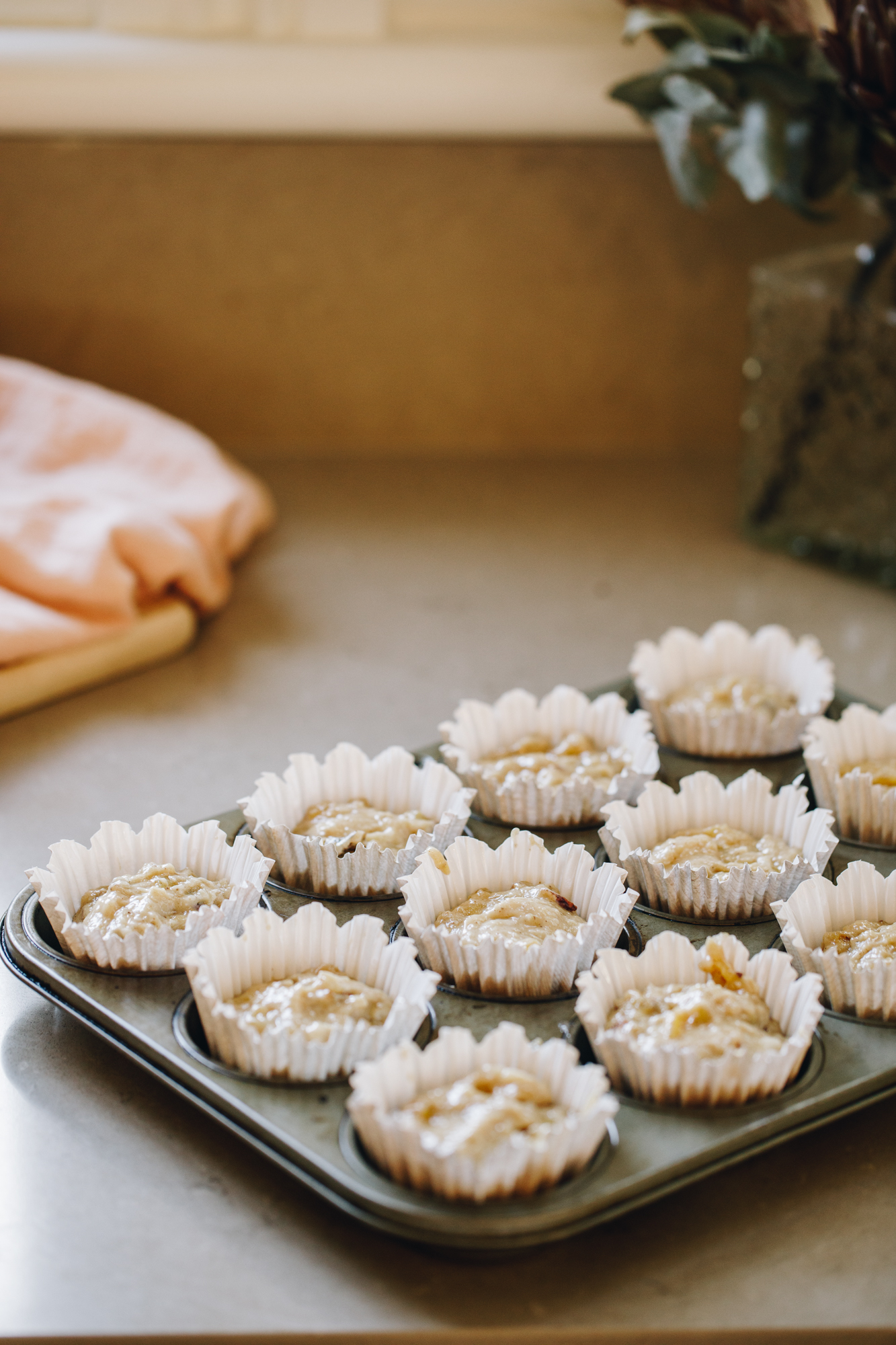 Muffin mixture in white muffin cases in a muffin tin, on a stone bench. 