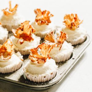 Cupcakes in white cases in a muffin tray, topped with dried pineapple flowers.