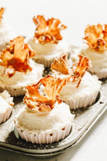 Cupcakes in white cases in a muffin tray, topped with dried pineapple flowers.