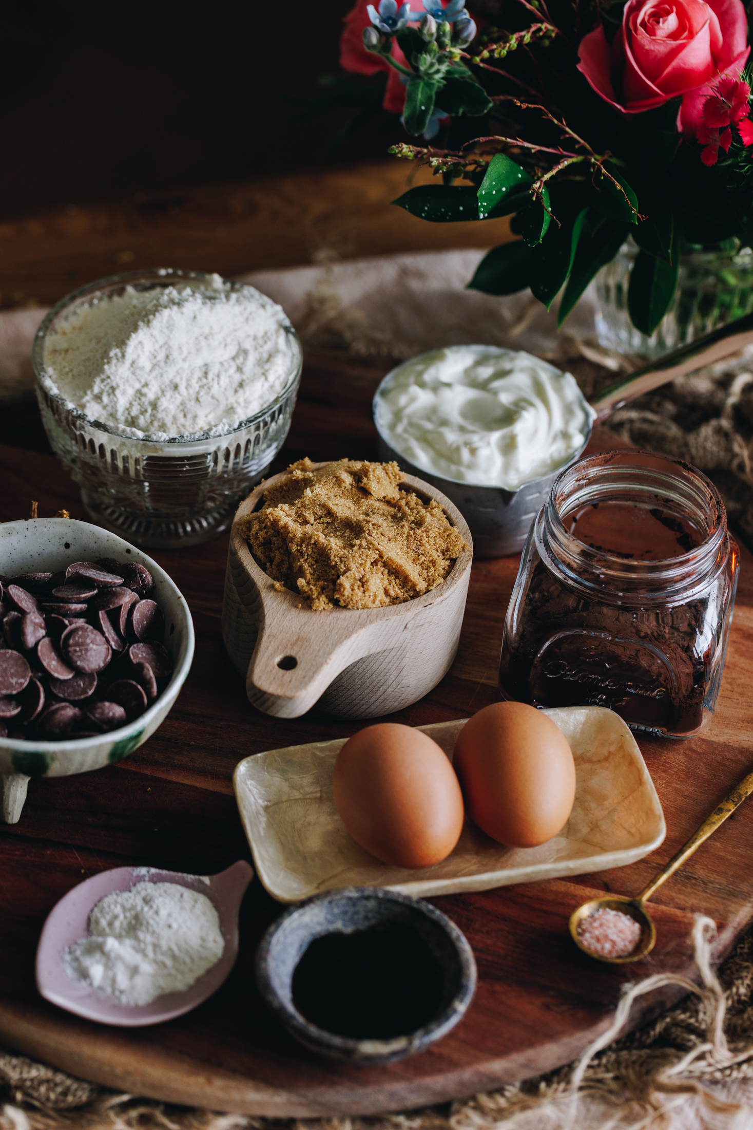 Chocolate muffin ingredients on a wooden board with pink flowers in the background. 