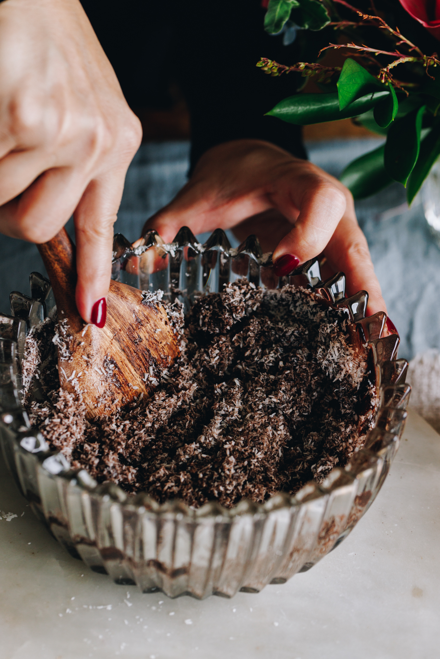 A glass vintage bowl with a cocoa and coconut mixture, stirred with a wooden spoon. Flowers in the background.  