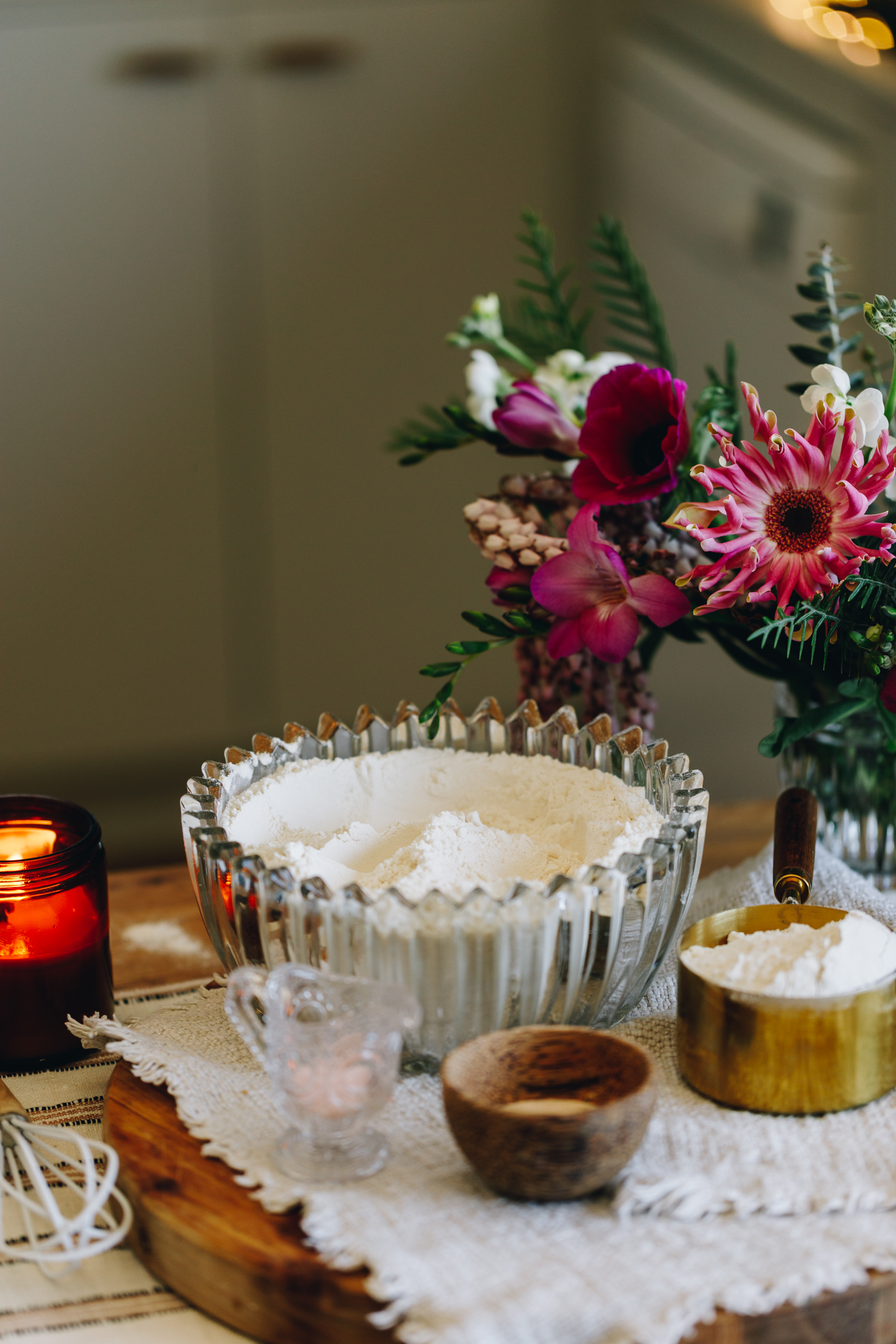 A wooden board with a glass bowl of flour and bagel ingredients on a white cloth, flowers in the background. 