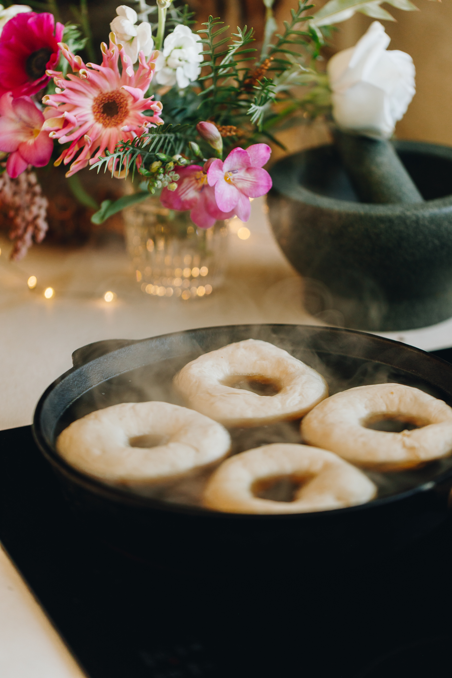 Bagels boiling in a cast iron pan with mortar and pestle and flowers in the background. 